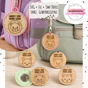 Könnte beinhalten: Runde Taschenanhänger aus Holz mit eingravierten Tiergesichtern und dem deutschen Text "Handy ran Mama dran" oder "Handy ran Papa dran". Die Anhänger haben rosafarbene oder hellgrüne kreisförmige Rahmen und Metallclips zur Befestigung an Rucksäcken.