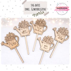 Könnte beinhalten: Holzdekorationen in Form einer Gießkanne mit Blumenarrangements und verschiedenen deutschen Sätzen. Die Dekorationen enthalten Sätze wie "Beste Mama" und "Alles Liebe zum Muttertag". Das Design ist lasergeschnitten und enthält einen langen Stab.