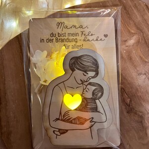 May include: A wooden card with a light-up heart and a drawing of a mother holding a baby. The card reads "Mama, du bist mein Fels in der Brandung - danke f&uuml;r alles!"