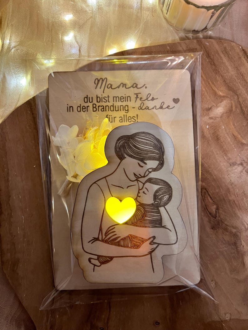 May include: A wooden card with a light-up heart and a drawing of a mother holding a baby. The card reads "Mama, du bist mein Fels in der Brandung - danke f&uuml;r alles!"