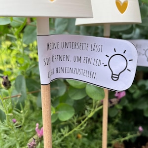 May include: White, heart-shaped garden lights with yellow heart cutouts. The lights are on wooden stakes, with a banner that reads, "Meine Unterseite l&auml;sst sich &ouml;ffnen, um ein LED-Licht hineinzustellen." The background is green foliage.