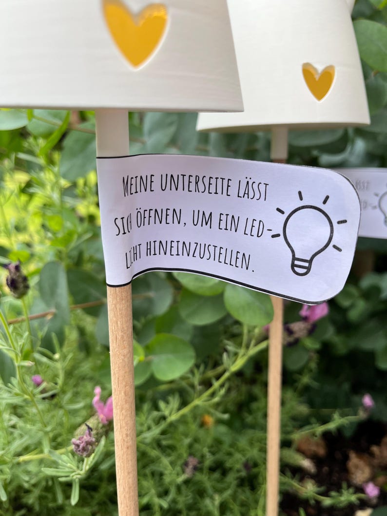 May include: White, heart-shaped garden lights with yellow heart cutouts. The lights are on wooden stakes, with a banner that reads, "Meine Unterseite l&auml;sst sich &ouml;ffnen, um ein LED-Licht hineinzustellen." The background is green foliage.