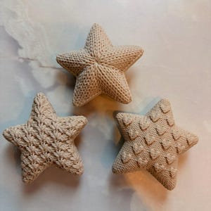 Puede incluir: Tres adornos en forma de estrella de color beige con texturas de punto distintas. Cada estrella presenta un patr&oacute;n &uacute;nico: uno con punto acanalado, otro con punto liso y otro con un dise&ntilde;o de punto en relieve tipo chevron.