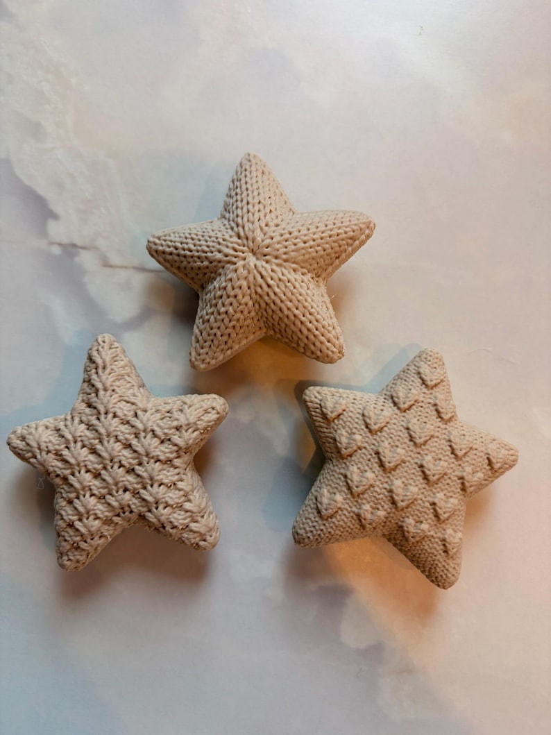 Puede incluir: Tres adornos en forma de estrella de color beige con texturas de punto distintas. Cada estrella presenta un patr&oacute;n &uacute;nico: uno con punto acanalado, otro con punto liso y otro con un dise&ntilde;o de punto en relieve tipo chevron.