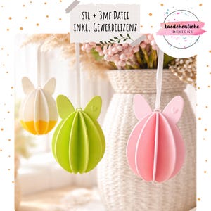 Puede incluir: Tres adornos colgantes de huevos de Pascua en blanco y amarillo, verde y rosa. Cada adorno tiene un diseño de orejas de conejo y está suspendido por una cinta blanca. El fondo incluye un jarrón con flores.