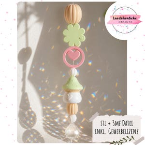 May include: A decorative hanging mobile with a pumpkin, clover, heart, mushroom, and crystal. The pastel-coloured mobile includes the text "Laedchenliebe Designs". The design incorporates a variety of shapes and colours.