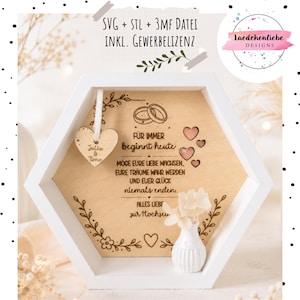 May include: A white hexagonal wooden shadow box frame containing a light wood panel engraved with German text, floral motifs, and two rings. A small wooden heart tag labeled Julia & Timo hangs inside, alongside a miniature white vase with flowers.