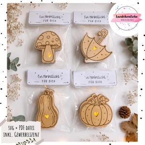 May include: Four clear plastic bags containing light-colored wooden cutouts in the shapes of a mushroom, an umbrella, and two pumpkins. Each cutout features a small heart-shaped cutout with a warm yellow light. Each bag has a white header card with the text "Ein Herbstlicht FÜR DICH".