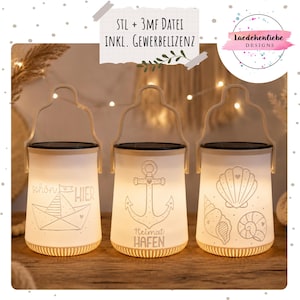 May include: Three white solar lanterns with cream-coloured handles and bases. Each lantern features a different nautical design: a sailboat, an anchor, and seashells. The lanterns have the text "schön HIER", "Heimat HAFEN", and the seller's logo.
