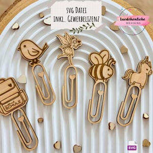 Può includere: Graffette di legno con vari disegni, tra cui un uccello, una fata, un'ape e un unicorno. Ogni graffetta presenta un dettaglio a forma di cuore ed è realizzata in legno chiaro. Le graffette sono disposte su una superficie bianca arcuata.