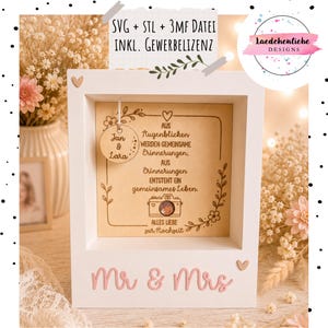 May include: A white wooden shadow box frame featuring the text 'Mr & Mrs' in pink script. Inside, a light wood panel displays German text, a camera illustration, and a hanging circular wooden tag engraved with 'Jan & Lara'.
