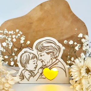 May include: A wooden cutout of an older woman and a child embracing, with a glowing yellow heart in the centre. The cutout is on a light brown wooden surface with white flowers.