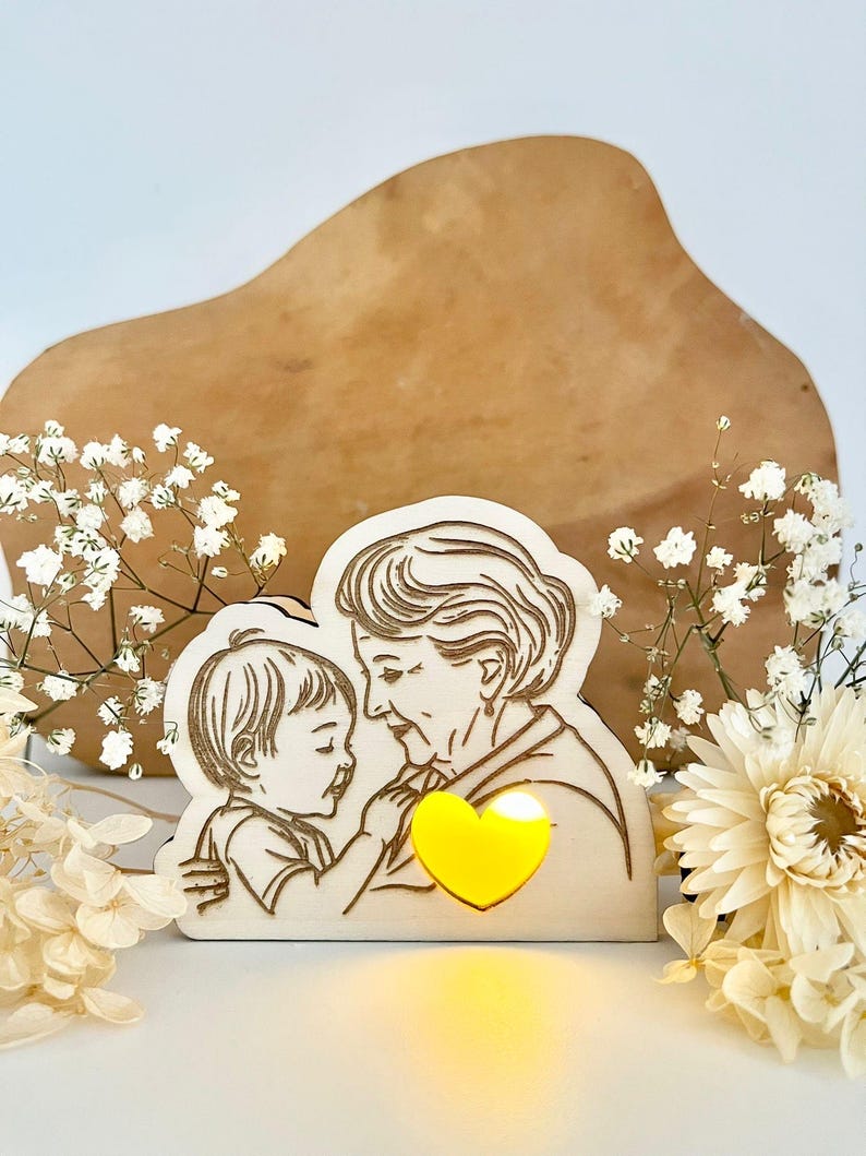 May include: A wooden cutout of an older woman and a child embracing, with a glowing yellow heart in the centre. The cutout is on a light brown wooden surface with white flowers.