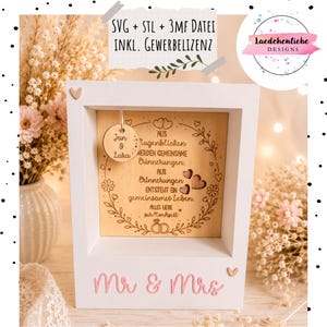 May include: A white wooden shadow box frame featuring a light wood panel with laser-engraved German text, a hanging circular wooden tag with the names Jan & Lara, and three small wooden hearts. The frame base displays the pink text Mr & Mrs.