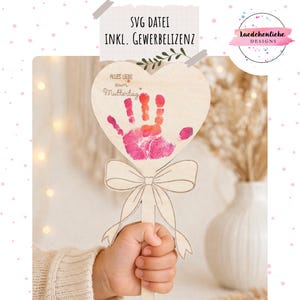 Könnte beinhalten: Ein herzförmiges Holzschild mit einem Handabdruck in Rosa und Orange und dem deutschen Text "Alles Liebe zum Muttertag". Das Schild hat auch eine Schleife und einen Griff. Der Text "SVG DATEI INKL. GEWERBELIZENZ" steht oben.
