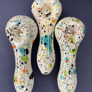 May include: Three speckled glass pipes with a cream-colored base and colorful paint splatters in black, teal, orange, and green. The pipes have a rounded bowl and a long, curved stem. The background is a solid dark gray.