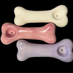 May include: Three ceramic pipes shaped like dog bones, arranged in a stack. The top pipe is off-white, the middle pipe is a glossy light pink, and the bottom pipe is a soft lavender. Each pipe features a circular bowl opening on one end.