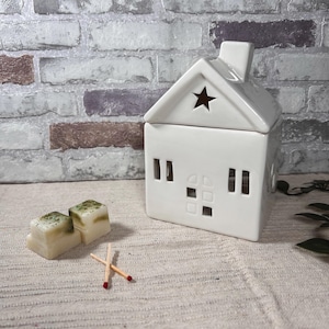 Könnte beinhalten: Ein weißer, hausförmiger Keramik-Duftlampenhalter mit Sternaussparung und rechteckigen Fenstern. Zwei quadratische Wachs-Schmelzstücke mit grünen Sprenkeln und zwei Streichhölzer befinden sich vor dem Wärmer. Der Hintergrund ist eine Backsteinmauer.