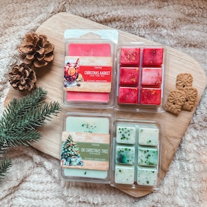 Puede incluir: Dos recipientes de plástico transparente llenos de cera aromática. El recipiente superior contiene cubos de cera roja con purpurina dorada y una etiqueta que dice Christmas Market. El inferior contiene cubos de cera verde claro con purpurina verde y una etiqueta que dice Oh Christmas Tree.