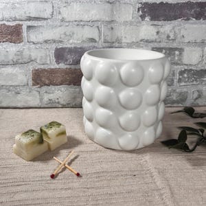 Può includere: Un portacandele in ceramica bianca con un design strutturato a forma di bolle. Due candele profumate quadrate con accenti verdi e blu sono posizionate accanto al portacandele, con due fiammiferi incrociati.