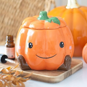 Può includere: Un contenitore in ceramica a forma di zucca con coperchio, con un volto sorridente, occhi neri e guance rosa. La zucca è arancione con uno stelo verde e piedi marroni. Si trova su una tavola di legno.