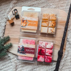 Op de afbeelding: Twee doorzichtige plastic houders voor waxmelts met labels waarop Buttery Beer en Wizard Express staat. De Buttery Beer wax is beige met bruine spikkels en de Wizard Express wax is wit met rode wervelingen en stippen.