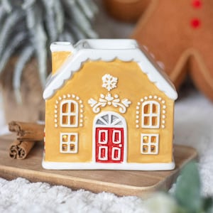 Könnte beinhalten: Ein Keramik-Lebkuchenhaus-Kerzenhalter mit gelber Außenseite, weißen Zuckergussdetails und einer roten Tür. Das Haus hat Fenster und einen Schornstein, ideal für die Weihnachtsdekoration. Es steht auf einem Holztablett mit Zimtstangen.