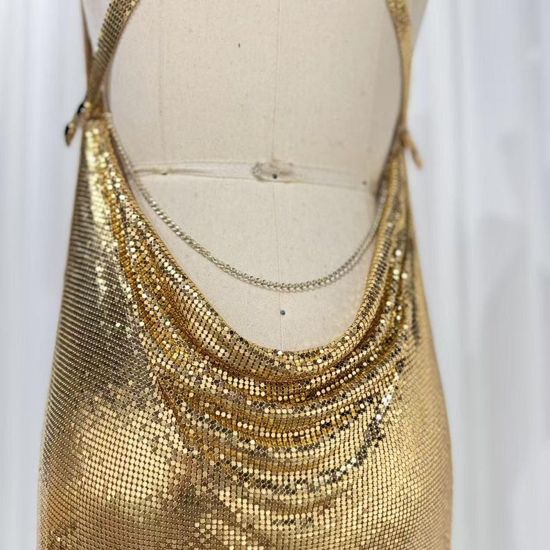 May include: A gold-toned metallic chainmail halter top featuring a draped cowl neckline and a thin, delicate gold-colored chain strap that secures across the back. The garment is constructed from small, interlocking metal links.