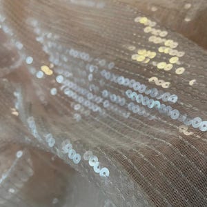 Könnte beinhalten: Nahaufnahme eines transparenten, hellpfirsichfarbenen Stoffes, der mit kleinen, runden, silbernen Pailletten bedeckt ist. Die Pailletten sind dicht gepackt und reflektieren das Licht, wodurch ein schimmernder Effekt entsteht. Der Stoff ist leicht zerknittert.