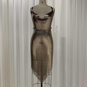 Puede incluir: Un vestido de malla dorada con cuello de pico y silueta ajustada. El vestido es sin mangas y tiene un escote alto.