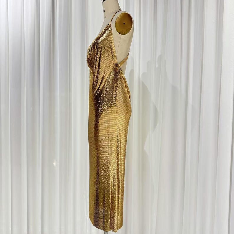 May include: A metallic gold chainmail midi dress featuring a cowl neckline, thin shoulder straps, and an open back design. The garment is displayed on a mannequin, showcasing a shimmering, textured surface that drapes along the length of the dress.