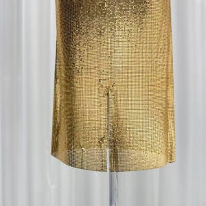 May include: A gold-colored metallic mesh garment featuring a fine, grid-like texture and a vertical slit at the bottom center. The material is composed of small, interlocking square links that create a shimmering, uniform surface across the entire piece.