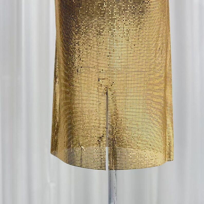 May include: A gold-colored metallic mesh garment featuring a fine, grid-like texture and a vertical slit at the bottom center. The material is composed of small, interlocking square links that create a shimmering, uniform surface across the entire piece.