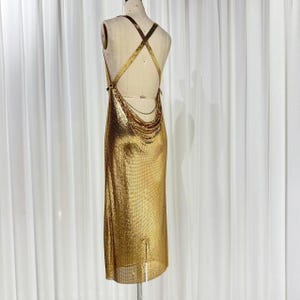 May include: A floor-length gold chainmail dress featuring a backless design with crisscross straps and a draped cowl detail at the lower back. The metallic fabric has a fine mesh texture that reflects light, creating a shimmering appearance.