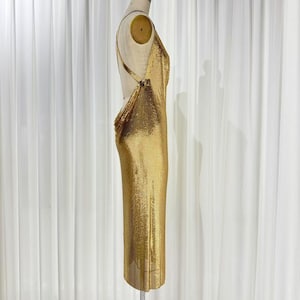 May include: A floor-length gold chainmail dress featuring a backless design with thin straps that connect at the sides. The metallic fabric has a fine, shimmering texture that drapes vertically along the form of the dress, creating a sleek silhouette.