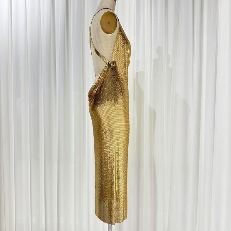 May include: A floor-length gold chainmail dress featuring a backless design with thin straps that connect at the sides. The metallic fabric has a fine, shimmering texture that drapes vertically along the form of the dress, creating a sleek silhouette.