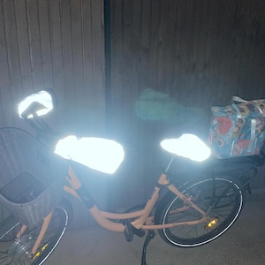 Può includere: Una bicicletta color pesca dotata di cestino anteriore grigio, coprimanubrio bianco riflettente, coprisella bianco riflettente e portapacchi posteriore con una borsa a motivo floreale nei toni del rosa e del blu, il tutto illuminato.