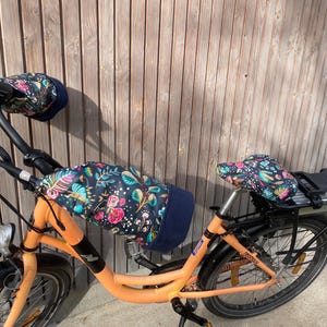 Könnte beinhalten: Ein orangefarbenes Fahrrad, ausgestattet mit einer dunkelblauen Lenkerabdeckung mit Blumenmuster, einem passenden Sitzbezug mit Blumenmuster und zwei dunkelblauen Lenkerstulpen mit Blumenmuster an den Griffen, mit rosa, gelben und grünen Blättern.