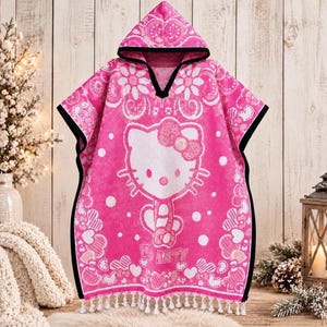 May include: A bright pink hooded poncho towel featuring a large white Hello Kitty graphic in the center. The fabric is decorated with white floral and heart patterns, finished with a black trim along the hood and edges, and white tassels at the hem.