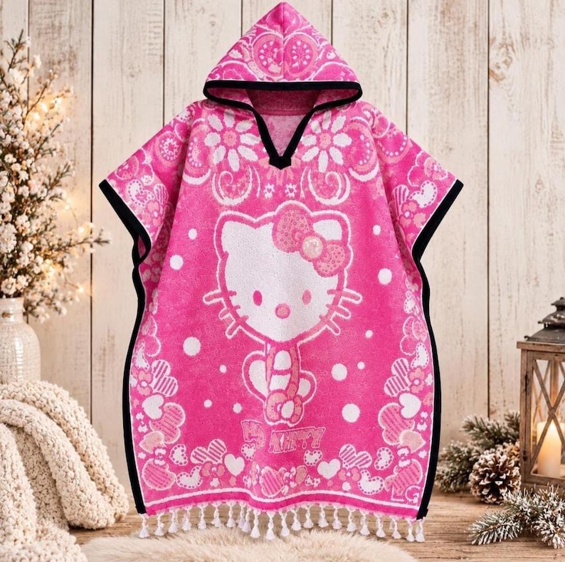 May include: A bright pink hooded poncho towel featuring a large white Hello Kitty graphic in the center. The fabric is decorated with white floral and heart patterns, finished with a black trim along the hood and edges, and white tassels at the hem.