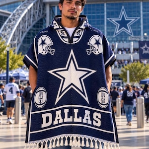 May include: A navy blue hooded poncho featuring a large white star graphic, two football helmet icons, and the word DALLAS printed in white block letters. The garment has a quarter-zip neckline and a white fringe trim along the bottom hem.
