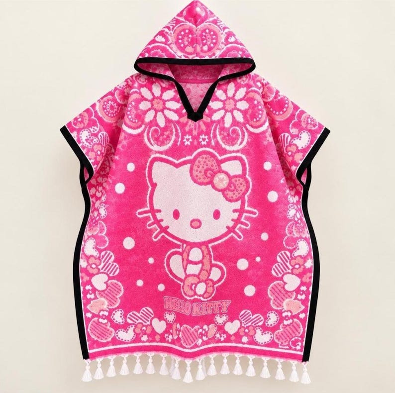 May include: A bright pink hooded poncho towel featuring a white Hello Kitty graphic, a bow, and the text Hello Kitty. The design includes floral and heart patterns with white polka dots, finished with a black trim and white fringe at the bottom.
