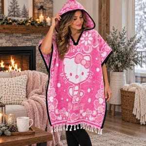 May include: A bright pink hooded poncho towel featuring a large white Hello Kitty graphic, a repeating floral and heart pattern, and black trim along the edges. The garment has a V-neckline and a hem finished with white fringe tassels.