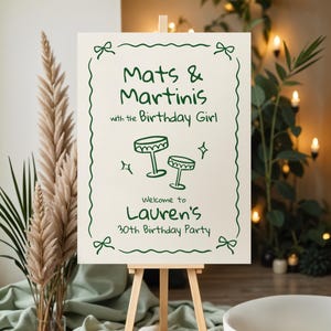 Pu&ograve; includere: Un cartello color crema su un cavalletto di legno con testo e illustrazioni verdi. Il cartello recita "Mats & Martinis with the Birthday Girl" e d&agrave; il benvenuto a "Lauren's 30th Birthday Party."