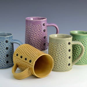 Könnte beinhalten: Fünf Keramikbecher mit einem Strickmuster und Knopfdetails. Die Becher sind in den Farben Lila, Blau, Grün und Gelb.