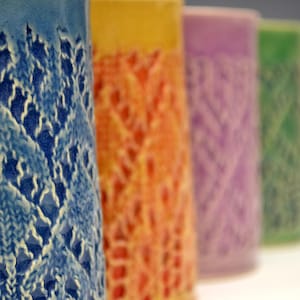 May include: A row of six ceramic mugs with a textured, lace-like design. The mugs are in shades of blue, orange, purple, green, yellow, and white.