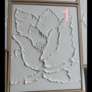 Op de afbeelding: Wit, getextureerd bloemenkunstwerk in een houten lijst. Het kunstwerk toont een grote bloem met gedetailleerde bloemblaadjes en een getextureerd oppervlak, waardoor een 3D-effect ontstaat. Het nummer "1" is zichtbaar. Het is een decoratief stuk.