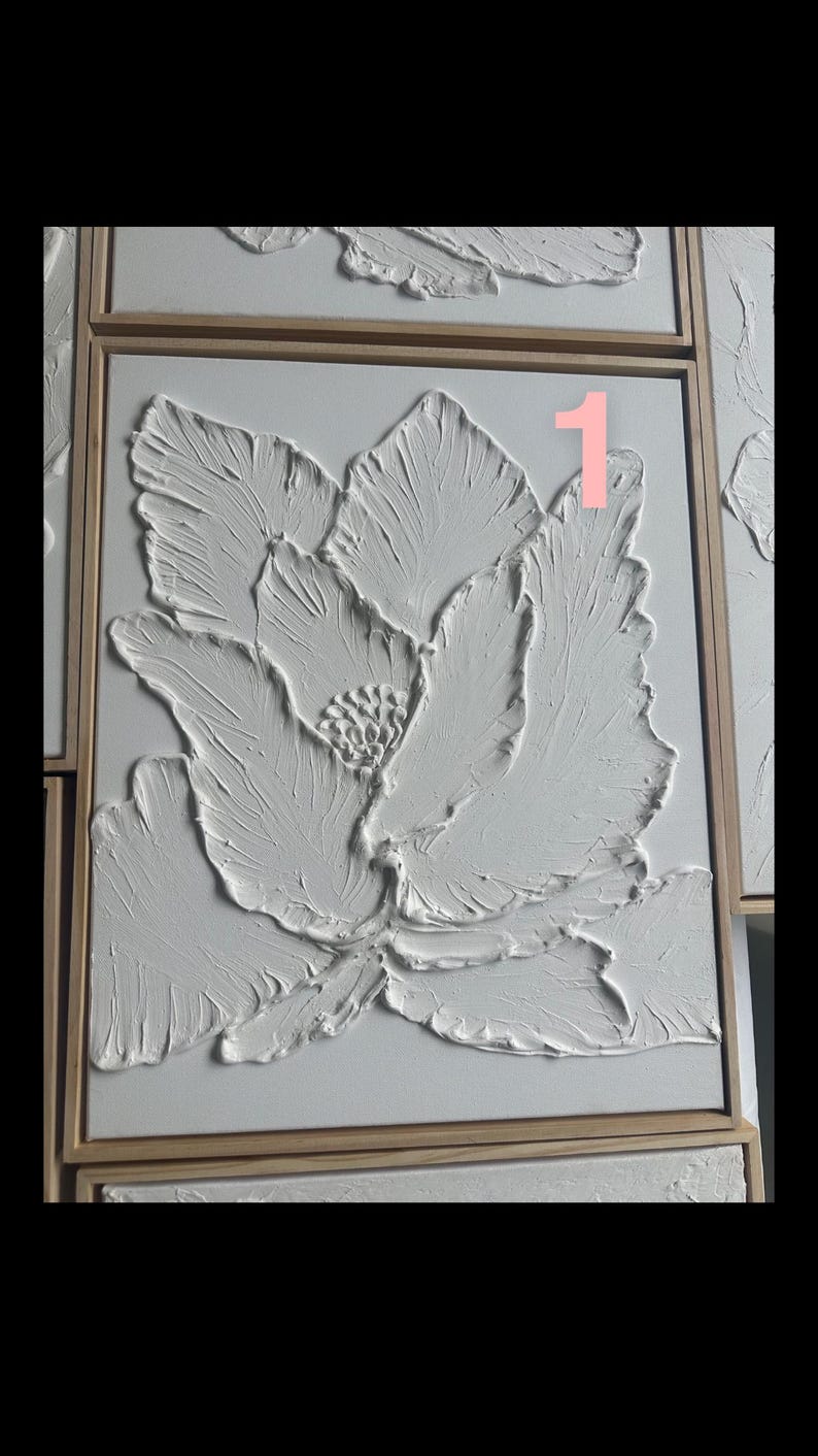 Op de afbeelding: Wit, getextureerd bloemenkunstwerk in een houten lijst. Het kunstwerk toont een grote bloem met gedetailleerde bloemblaadjes en een getextureerd oppervlak, waardoor een 3D-effect ontstaat. Het nummer "1" is zichtbaar. Het is een decoratief stuk.