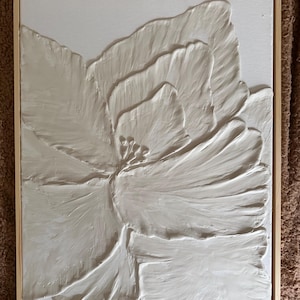 May include: A white, textured floral art piece in a natural wood frame. The artwork features a large, stylized flower with raised petals, creating a 3D effect. The neutral color palette and simple frame make it suitable for various decor styles.