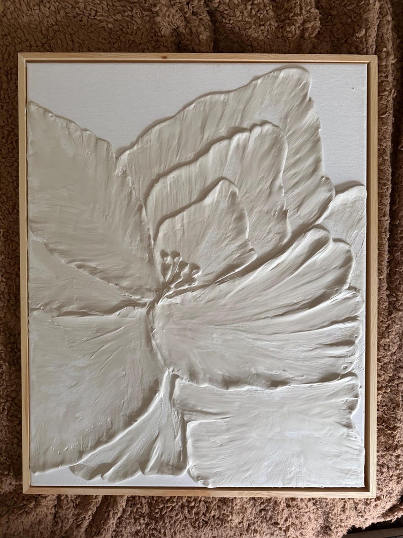 May include: A white, textured floral art piece in a natural wood frame. The artwork features a large, stylized flower with raised petals, creating a 3D effect. The neutral color palette and simple frame make it suitable for various decor styles.